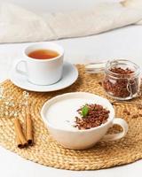 iogurte com granola de chocolate na xícara, café da manhã com chá em fundo bege, vista lateral, vertical. foto
