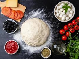 massa crua para pizza com ingredientes e especiarias em fundo preto escuro, vista superior, configuração plana foto