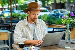 jovem hipster elegante no trabalho de chapéu com laptop ao ar livre no café de rua foto