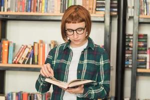 mulher jovem ruiva de óculos ler livro na biblioteca foto