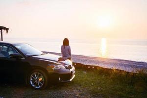 mulher fica ao lado do carro de luxo preto e olha para a vista panorâmica do mar calmo e do sol poente. tsikhisdziri. Geórgia. conceito de viagem por estrada e relaxamento solo pelo conceito de mar. foto