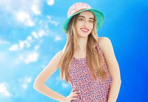 retrato de uma mulher atraente sorridente no vestido de verão e chapéu posando em pé e olhando para câmera isolada sobre fundo azul foto