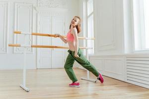 Motivada alegre mulher ruiva de sutiã esportivo e calça, mantém a mão no bolso, usa tênis, faz exercícios na barra de balé, dança no estúdio, está de bom humor, termina o treino de dança foto