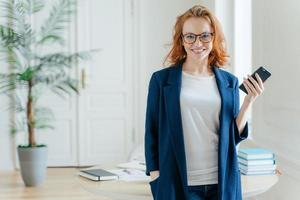 foto de meio comprimento de mulher bonita usa celular moderno para blogar em redes sociais, posa no interior do escritório, vestida com roupa formal, verifica a notificação, usa óculos ópticos tem cabelo ruivo