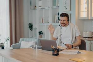 feliz freelancer masculino usando fone de ouvido e trabalhando em casa durante a quarentena enquanto olha para a tela do laptop