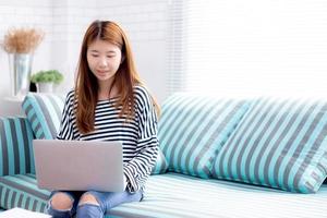 bela do retrato jovem mulher asiática usando laptop para lazer no sofá na sala de estar, garota trabalhando online com notebook freelance com um conceito de negócio de comunicação feliz. foto