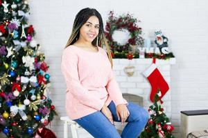 jovem e bela mulher afro-americana sorridente na camisola rosa está esperando o natal em casa. de pé no fundo de guirlandas e véspera de ano novo. foto