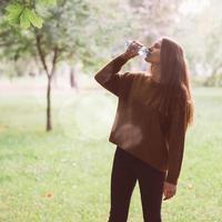 linda jovem bebendo água de uma garrafa de plástico na rua no parque no outono ou inverno. uma mulher com lindos cabelos escuros longos e grossos sacia sua sede de água em uma caminhada