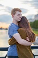 menina com escuro longo e grosso ouve abraçando menino ruivo de camiseta azul na ponte, amor adolescente ao pôr do sol foto