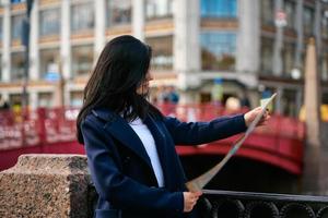 jovem de pé à beira-mar da cidade grande e olhando para o guia, turista à procura de atrações. uma encantadora mulher pensativa elegantemente vestida com longos cabelos escuros viaja pela europa foto