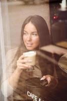 uma linda garota senta-se no café e olha pela janela pensativamente. reflexo da cidade na janela. mulher morena com cabelo comprido bebe café cappuccino, vertical