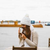 linda jovem bebendo café, chá de caneca plástica no outono, inverno. uma mulher com cabelos compridos em roupas quentes, sentado à beira-mar no mar Báltico no porto, aquecido por bebida quente