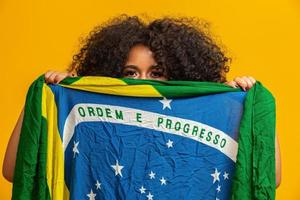 fã de mulher negra misteriosa segurando uma bandeira brasileira na sua cara. cores do brasil em fundo, verde, azul e amarelo. eleições, futebol ou política. foto