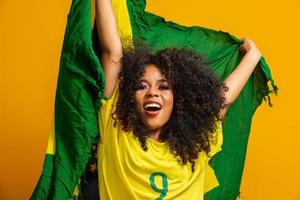 garota afro torcendo pelo time brasileiro favorito, segurando a bandeira nacional em fundo amarelo. foto