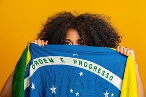 fã de mulher negra misteriosa segurando uma bandeira brasileira na sua cara. cores do brasil em fundo, verde, azul e amarelo. eleições, futebol ou política. foto