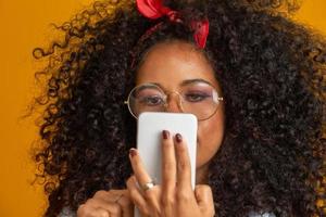 foto de estúdio de entretida e feliz garota afro-americana com penteado afro segurando smartphone usando o dispositivo para se divertir. fundo amarelo.