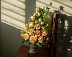 buquê de rosas artificiais falsas em um vaso em uma cadeira de madeira com luz solar persiana. foto