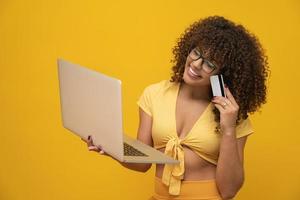 jovem linda garota de cabelo encaracolado com laptop e cartão de crédito em fundo amarelo, mulher de negócios compras online. foto