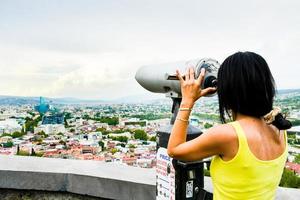 jovem mulher caucasiana fica no ponto de vista, aprecia o panorama através de binóculos na famosa fortaleza de narikala, capital da geórgia, tbilisi, na primavera foto