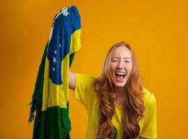 torcedor brasileiro. fã de mulher ruiva brasileira comemorando no futebol, partida de futebol em fundo amarelo. cores do brasil. vestindo camiseta, bandeira e chapéu de torcedor.