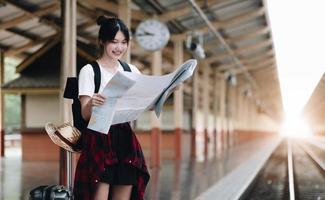 jovem viajante com bagagem e chapéu olhando para o mapa com fundo de trem na estação de trem. conceito de viagem foto