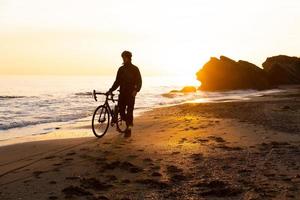 silhueta de jovem ciclista masculino no capacete na praia durante o belo pôr do sol foto