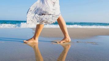 jovem mulher bonita vestida com um vestido branco anda descalço na praia de verão foto