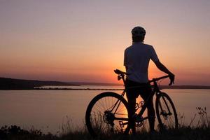 silhueta de um ciclista assistindo o pôr do sol no lago, ciclista masculino no capacete durante o pôr do sol foto
