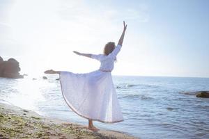 jovem caminhando na praia de manhã em lindo vestido branco. apto feminino se divertindo durante o nascer do sol. foto