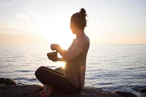 jovem esportiva jogar na tigela tibetana e sentado nas pedras, linda mulher asiática assistindo o nascer do sol no mar em pose de ioga foto