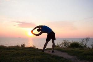 corredor de ajuste feliz assistindo o nascer ou o pôr do sol com punhos erguidos, jovem atleta na grama durante o nascer do sol no mar foto