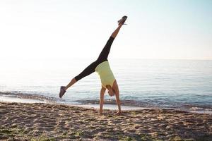 jovem ginasta profissional dança na praia, exercícios de treinamento com saltos legais, nascer do sol no fundo do mar ou oceano foto