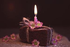 bolo de brownie de chocolate com nozes com vela de aniversário única com chama apagada com conjunto de fumaça fluindo e pano de fundo escuro com flores roxas. copie o espaço.