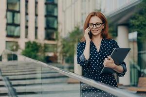 mulher ruiva em roupas da moda tem conversa telefônica anda na rua perto de prédio moderno foto