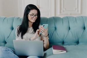 freelancer morena bem sucedida assiste a transmissão ao vivo de vídeo online, posa no sofá confortável com aparelhos modernos, lê mensagem no celular, trabalha distante de casa, vestido casualmente foto