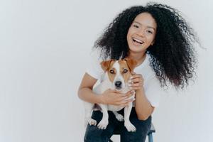 retrato de alegre garota encaracolada acariciando seu cachorro, regozijando-se comprando jack russell terrier, sorri amplamente, brinca com animais, usa roupas casuais, isoladas sobre fundo branco, goza de bom dia