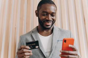 feliz empresário africano de terno segurando cartão de crédito e celular enquanto faz compras online foto