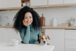 pessoas, animais, conceito de amizade. alegre mulher encaracolada senta-se na cozinha, bebe bebidas quentes, seu leal animal de estimação posa perto de desfrutar de passar tempo juntos. garota afro com cachorro em apartamento moderno foto