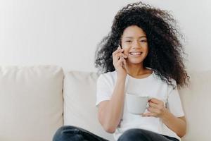 foto horizontal de mulher bonita tem conversa distante via celular, resolve questões informais enquanto fala, senta-se no sofá na sala, bebe chá, tem sorriso largo, mostra dentes brancos