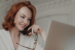 profissional sorridente web designer feminina de gengibre usa aplicativo no laptop para criar trabalho de projeto, faz pesquisas on-line e navega na internet, segura óculos, tem expressão feliz foto