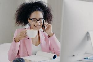 foto de empresária sorridente atraente gosta de bebida aromática, segura caneca branca de bebida, conversa por telefone, vestida com roupa formal, posa no local de trabalho, usa óculos transparentes