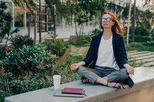 mulher de negócios pacífica de mãos dadas em gesto de mudra, sentado em pose de lótus e meditando no parque foto