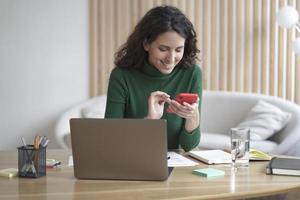 jovem mulher italiana feliz freelancer sentado no local de trabalho com laptop em casa e usando telefone celular foto