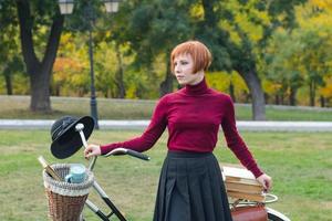 jovem no parque de outono leu livro, linda mulher ruiva com bicicleta na grama verde foto