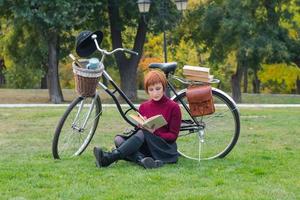 jovem no parque de outono leu livro, linda mulher ruiva com bicicleta na grama verde foto