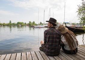 jovem casal de viajantes sentado no cais de madeira perto do rio, dois descolados com chapéus e dreadlocks de férias foto