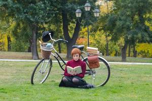 jovem no parque de outono leu livro, linda mulher ruiva com bicicleta na grama verde foto