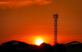 torre de telecomunicações com céu vermelho por do sol e nuvens. poste de rádio e satélite. tecnologia de comunicação. indústria de telecomunicações. rede 4g móvel ou de telecomunicações. silhueta casa rural e floresta. foto