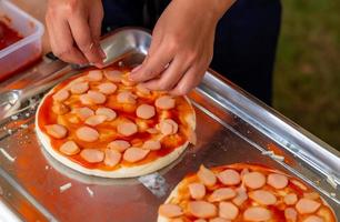 homem faz pizza na cozinha com massa de pizza e salsicha fatiada. pizza de dois pedaços na bandeja de aço inoxidável. conceito de fast food e junk food. alimentos não saudáveis. foto