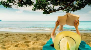 mulher deitar-se na toalha verde que colocou na praia de areia debaixo da árvore e lendo um livro. vida lenta nas férias de verão. mulher asiática com chapéu relaxante e curtindo férias na praia tropical. Ritmo de verão foto